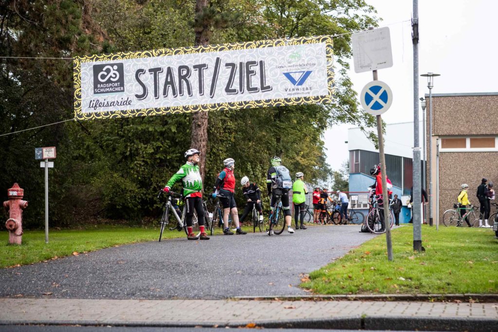 Start- und Zielbereich einer Radtourenfahrt mit Banner über der Straße, mehrere Radfahrer:innen stehen mit ihren Rädern bereit.