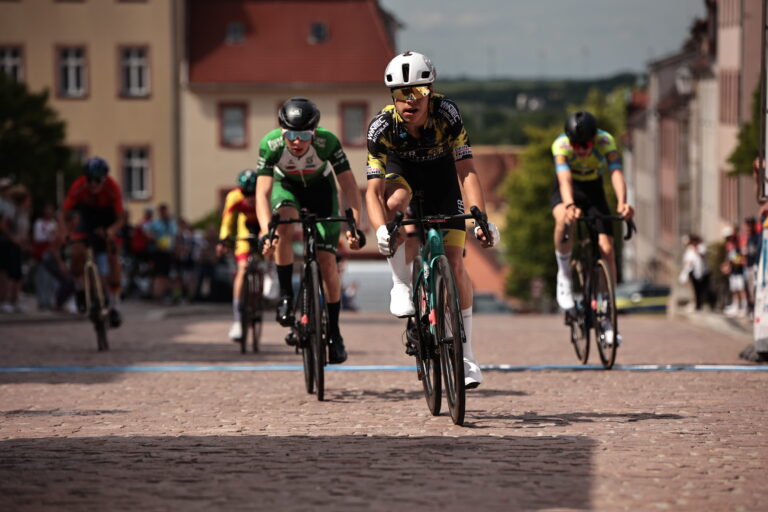 Radsportler:innen beim Zieleinlauf auf Kopfsteinpflaster mit Zuschauer:innen im Hintergrund, Fokus auf Fahrerin im VfR Büttgen Trikot.